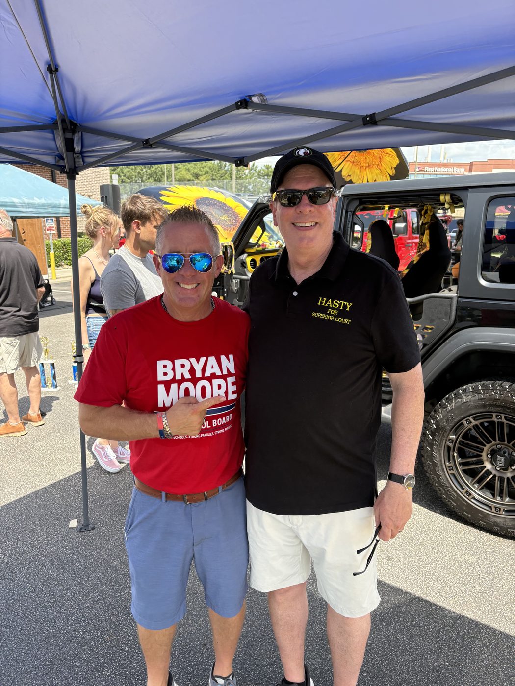 Bryan Moore in a campaign T-shirt under a tent at an outdoor community event, beside a man in a Hasty for Superior Court polo.