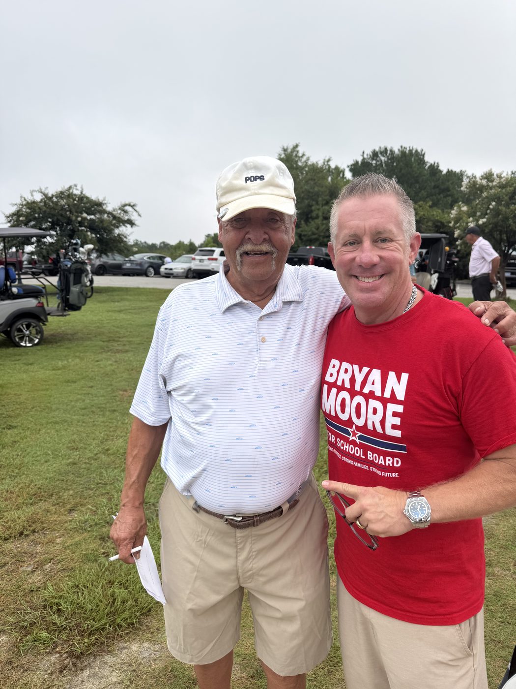 Bryan Moore in a campaign T-shirt with an older man in a golf cap, carts and bags behind them.