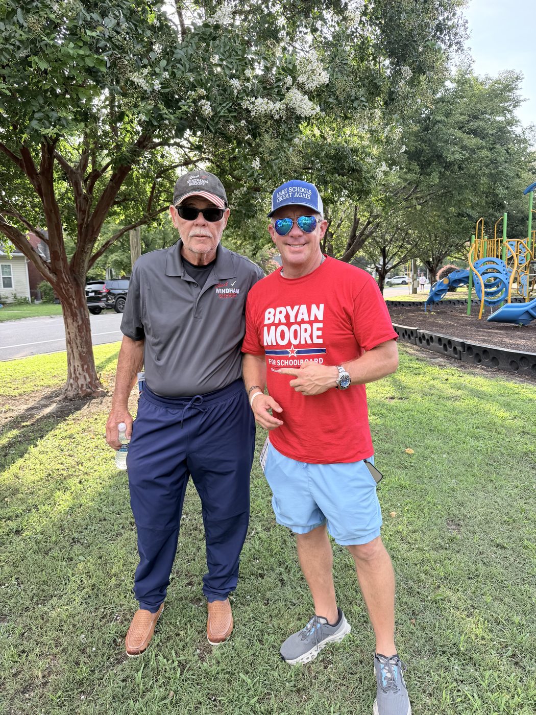 Bryan Moore in a campaign T-shirt and a cap reading Make Schools Great Again, standing beside a neighborhood playground with a man in Windham for Sheriff apparel.