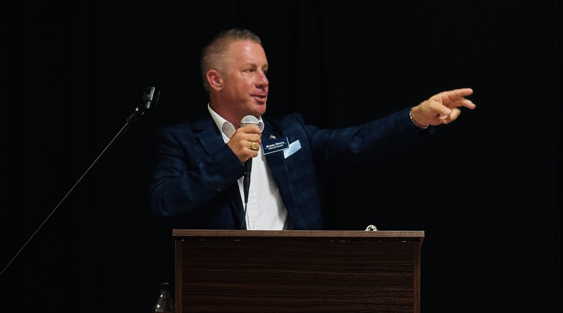 Bryan Moore speaking into a microphone at a lectern, one arm raised toward the audience.